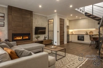 Современная уютная гостиная в нейтральной палитре: большой светло‑бежевый диван с подушками, деревянный каминный портал с длинным горизонтальным электрическим камином и настенным телевизором, акцентные картины и напольная лампа, кофейный столик на ковре, тёплый коричневый деревянный пол, справа открытая кухня в светлых латте‑тонах с подсветкой фартука и обеденным столом, на заднем плане лестница с деревянными ступенями и тёмными металлическими перилами — единое гармоничное пространство.