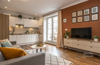 Стильный светлый интерьер квартиры с открытой планировкой: уютная гостиная с серым диваном и журнальным столиком на ковре, напротив — тумба с телевизором у карамельной акцентной стены с декоративными картинками; в глубине — современный кухонный уголок с белыми фасадами, деревянной столешницей и подсветкой, деревянный пол и большое окно во всю высоту, впускающее много естественного света.